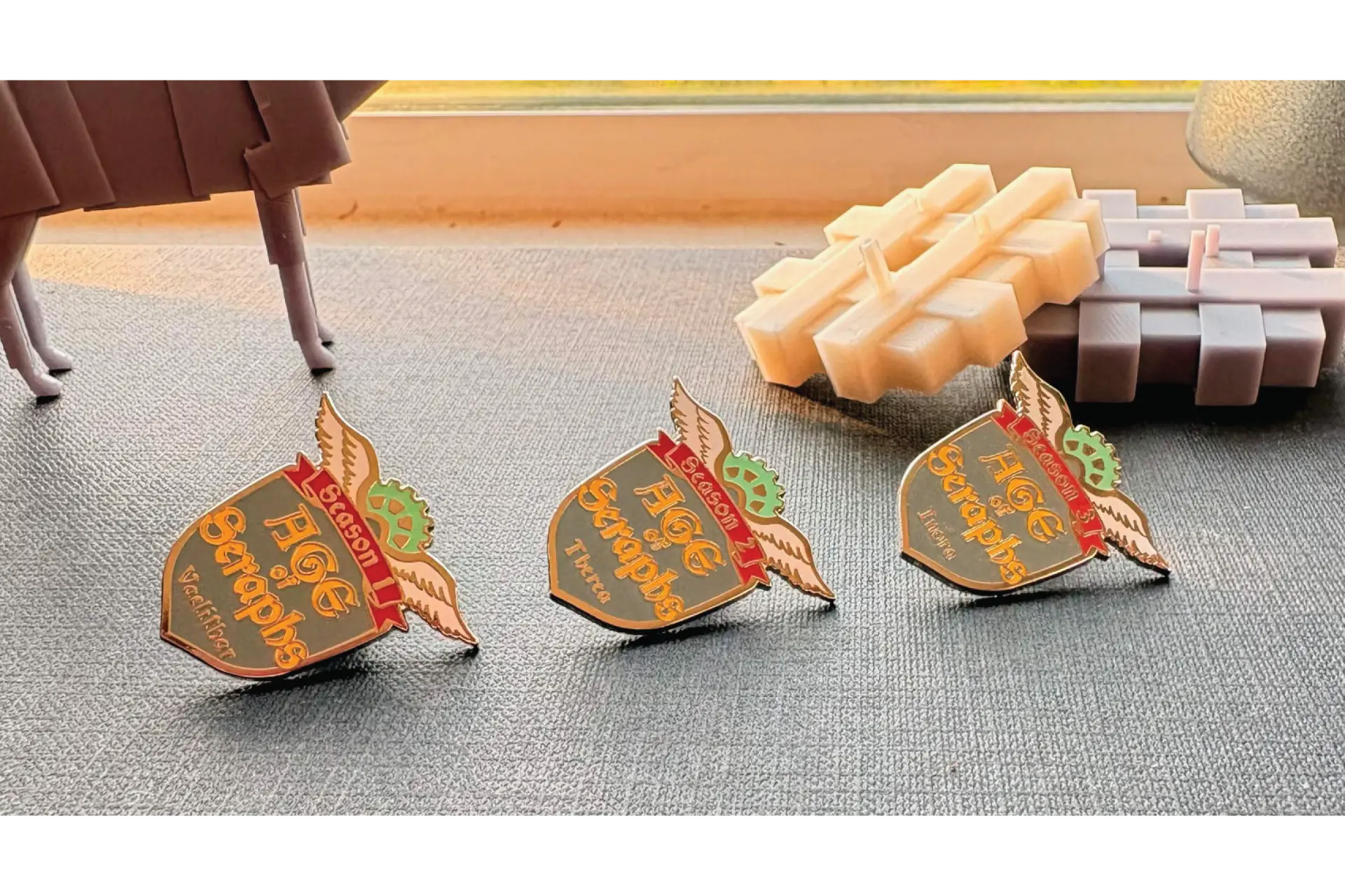 Age of Seraphs enamel pins staged with tabletop game terrain in warm window light