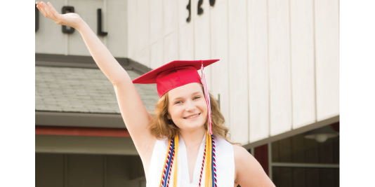 Graduate in a red cap and gown raising one arm outside a school building