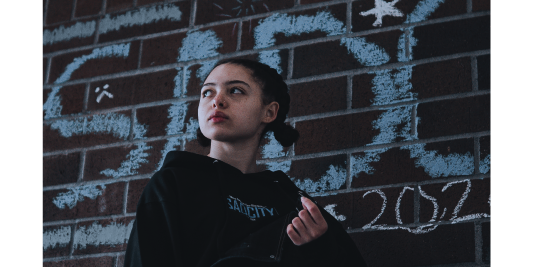 Young person in a black branded hoodie photographed against a brick wall with chalk lettering