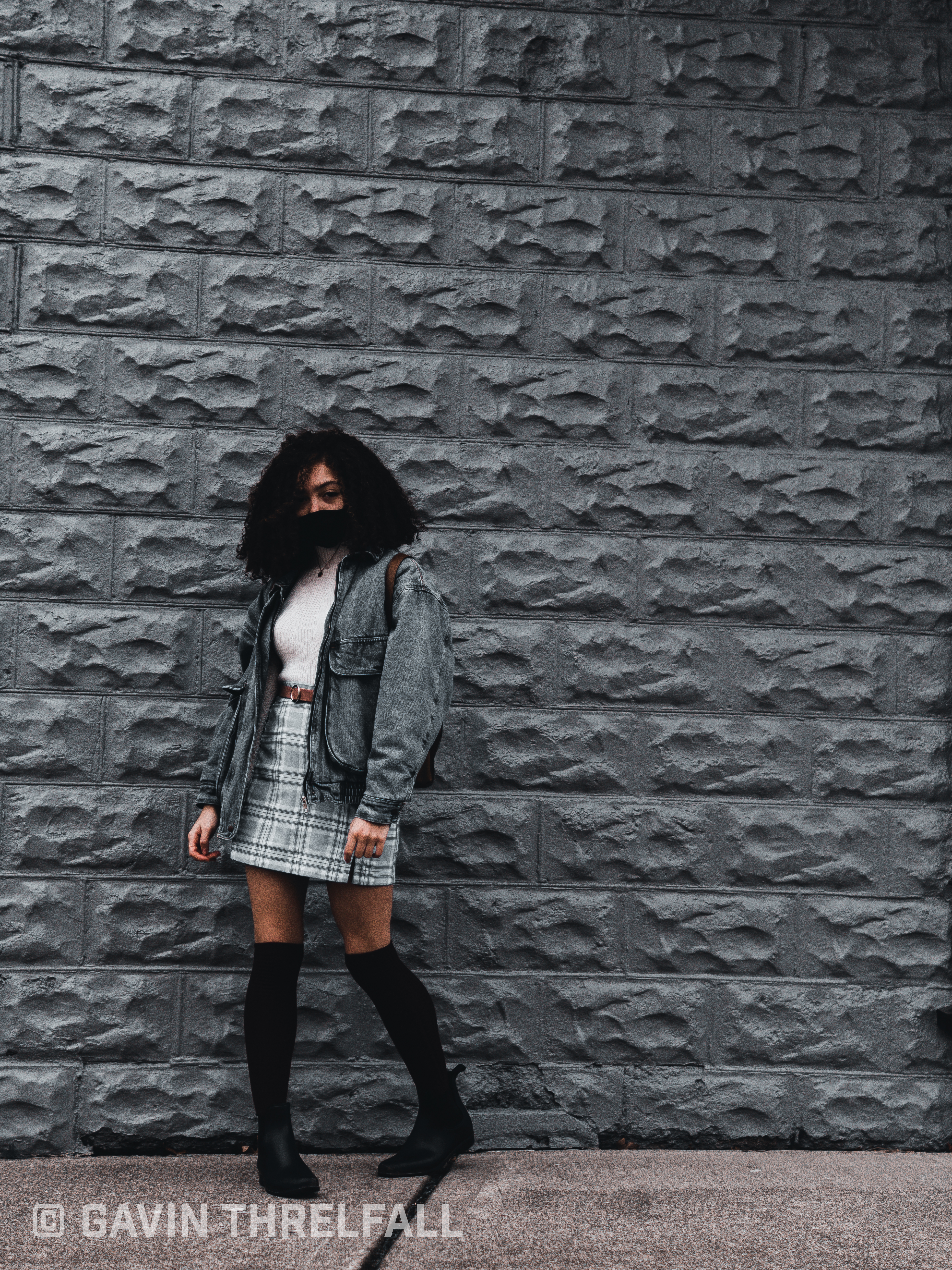 Model in denim jacket and plaid skirt posing against a grey brick wall