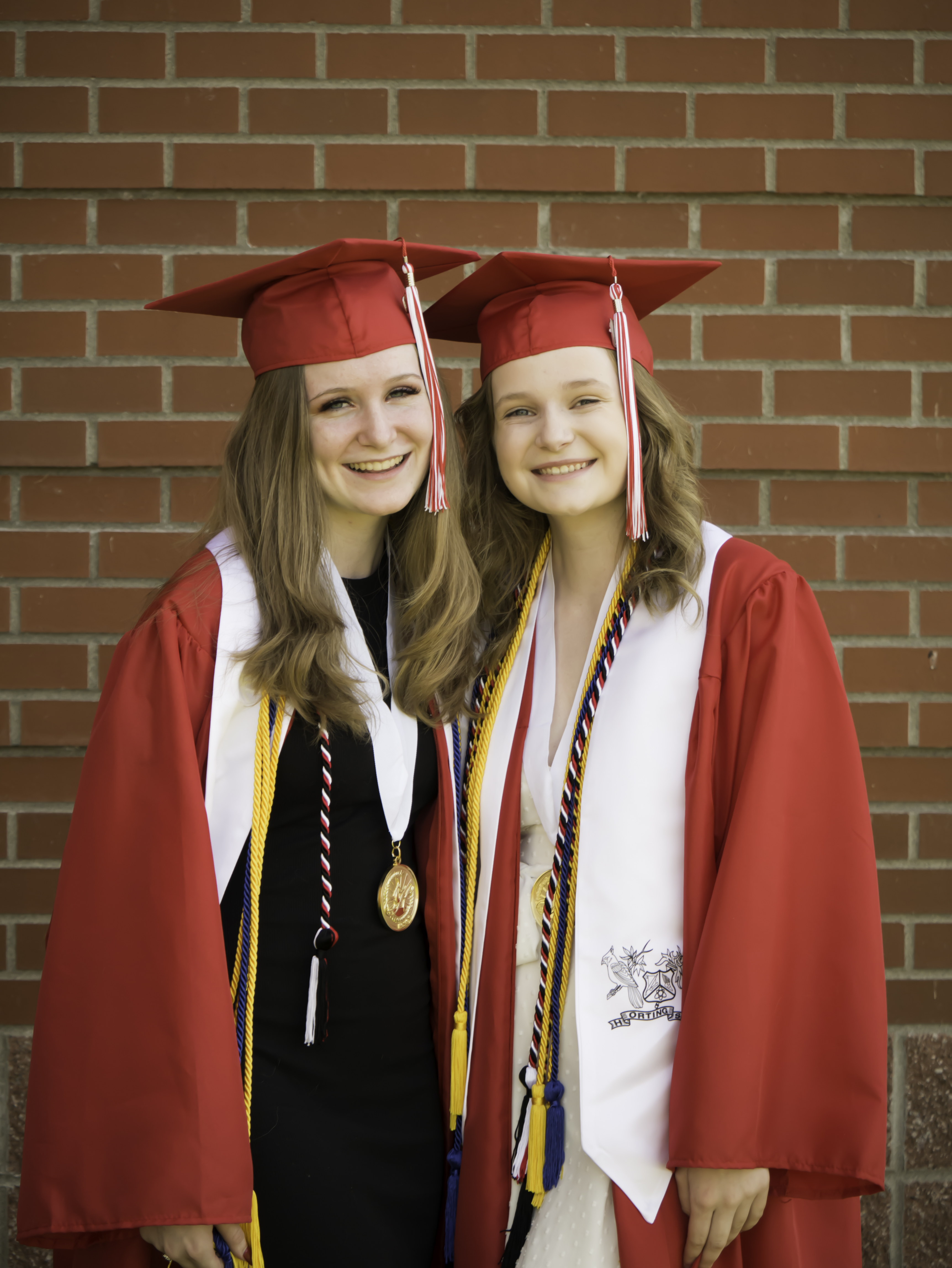 Twin sisters in red graduation caps and gowns beside a brick wall