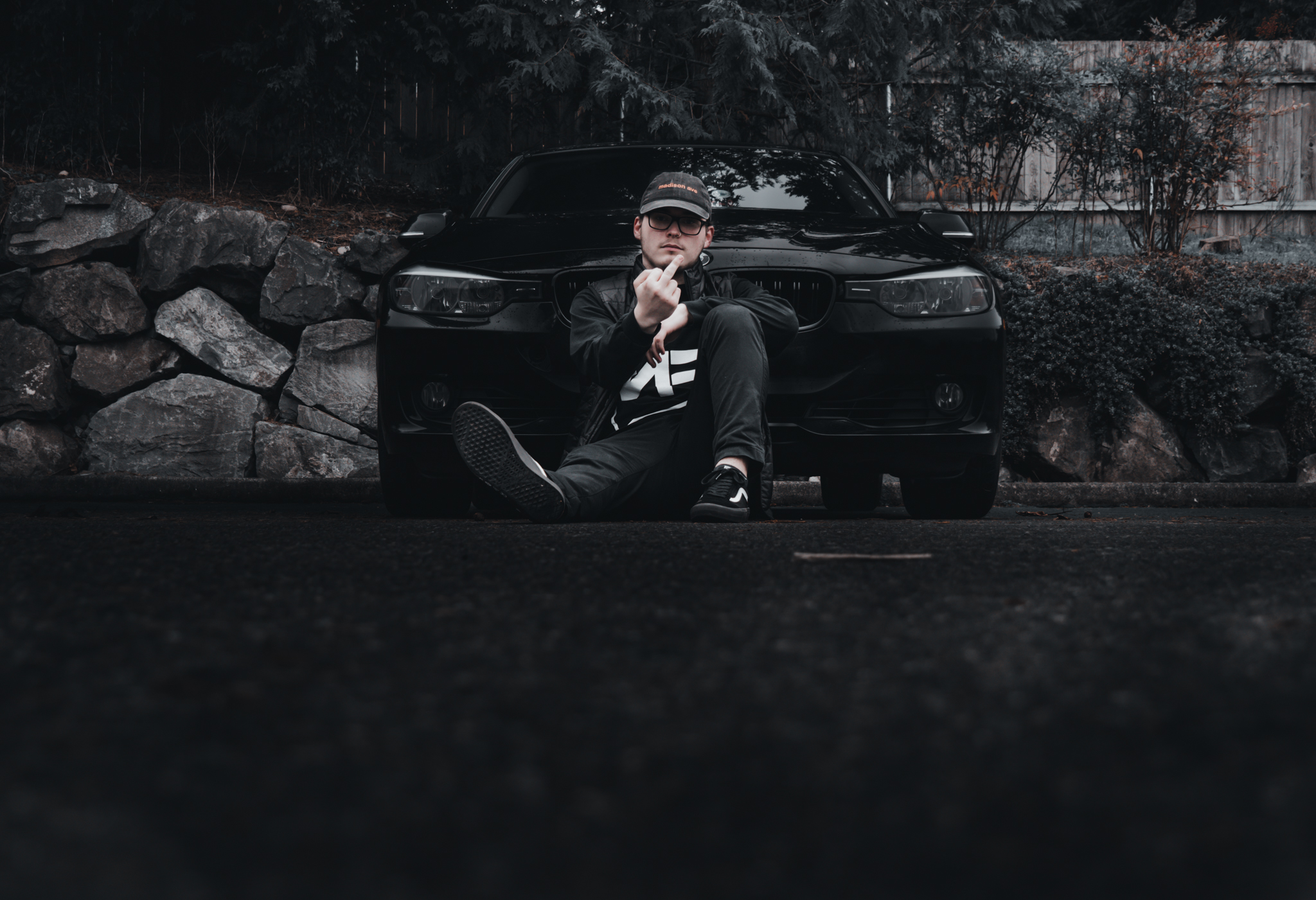 Man seated on pavement leaning against a black sedan on a rainy road