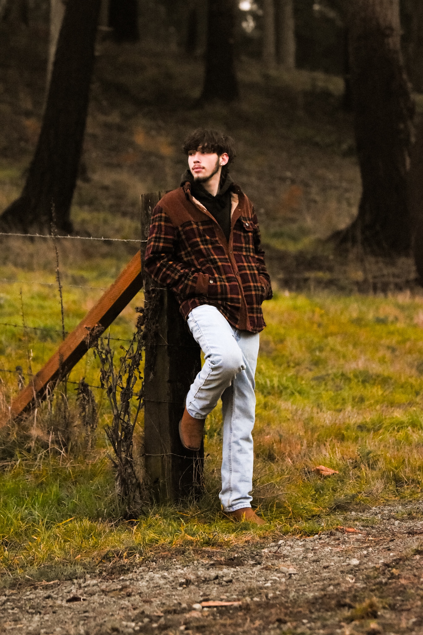 Young man in a plaid jacket leaning against a wooden fence post