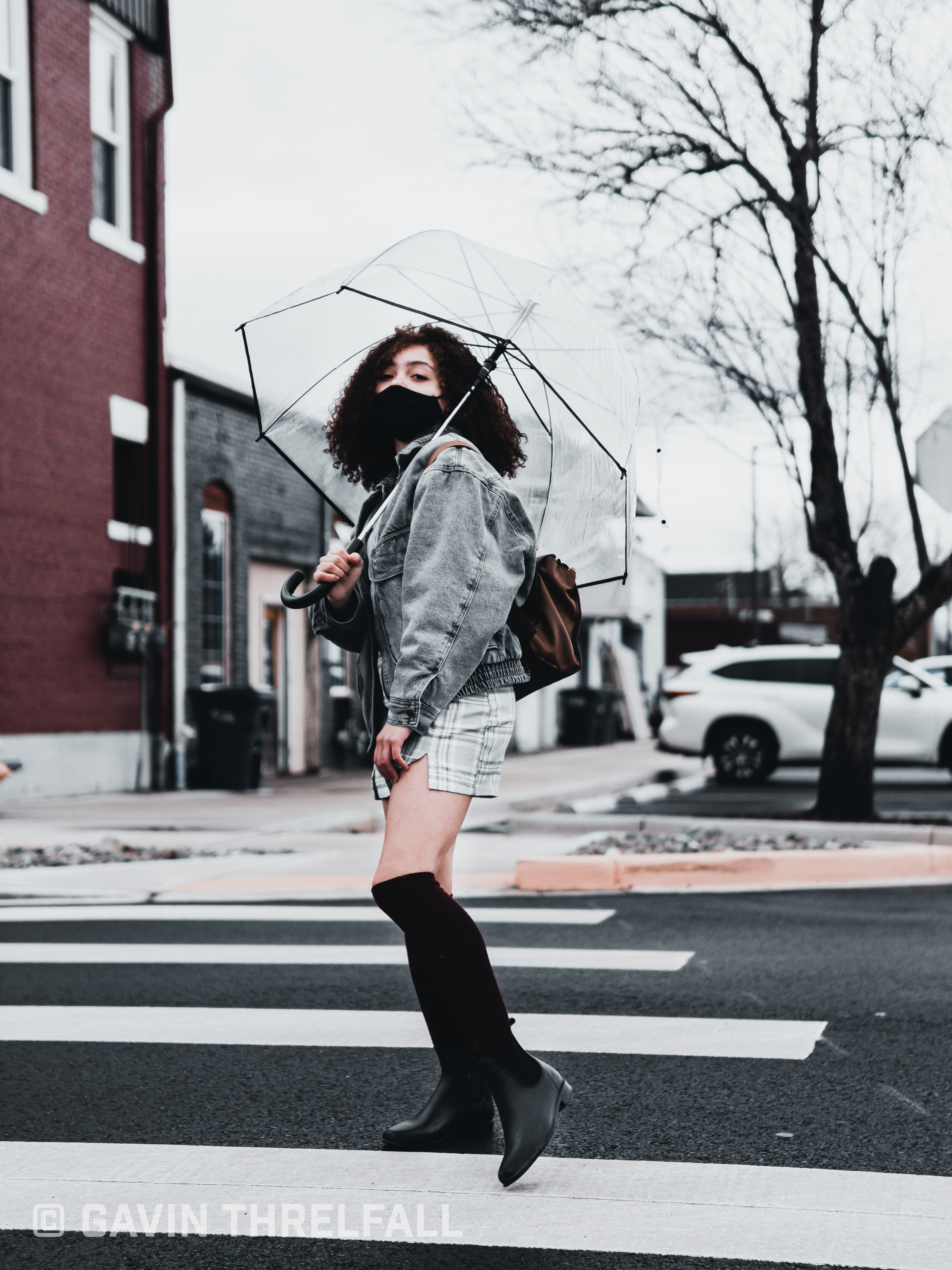 Model with a clear umbrella walking a crosswalk beneath bare trees